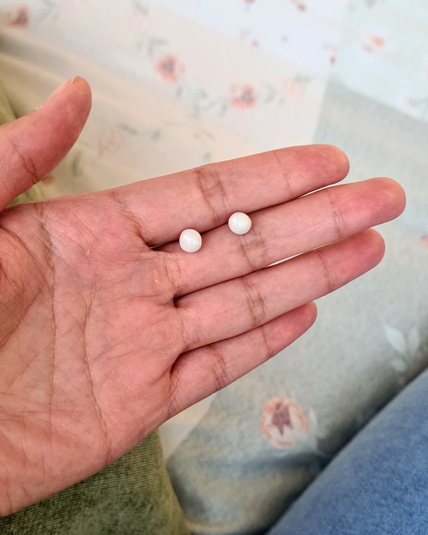 Two faux pearl studs held in a person's hand on a patterned fabric background