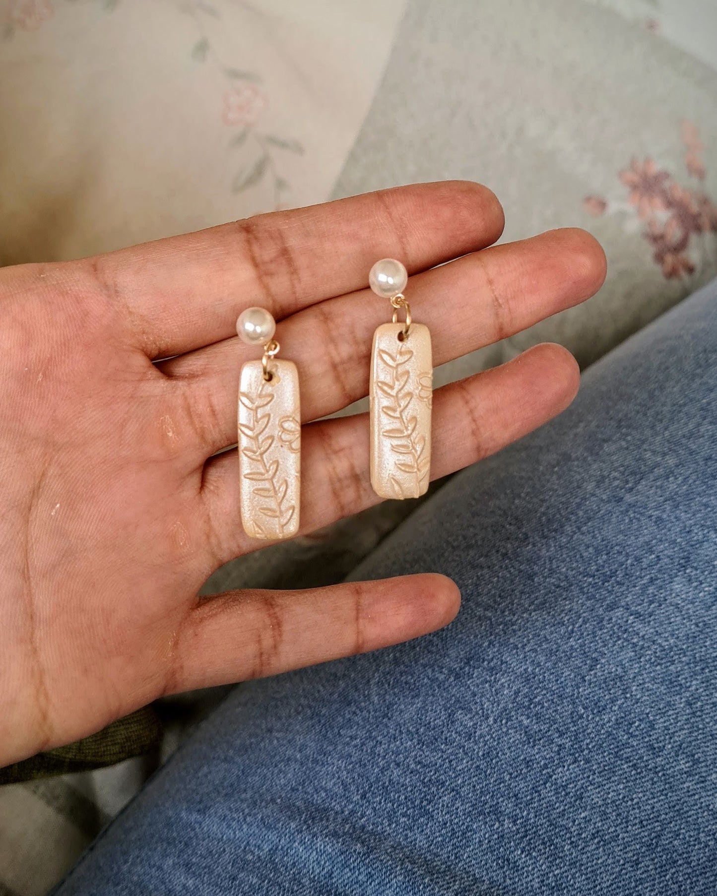 Hand holding shimmery yellow earrings with floral texture and pearl pendants on a floral-patterned surface.