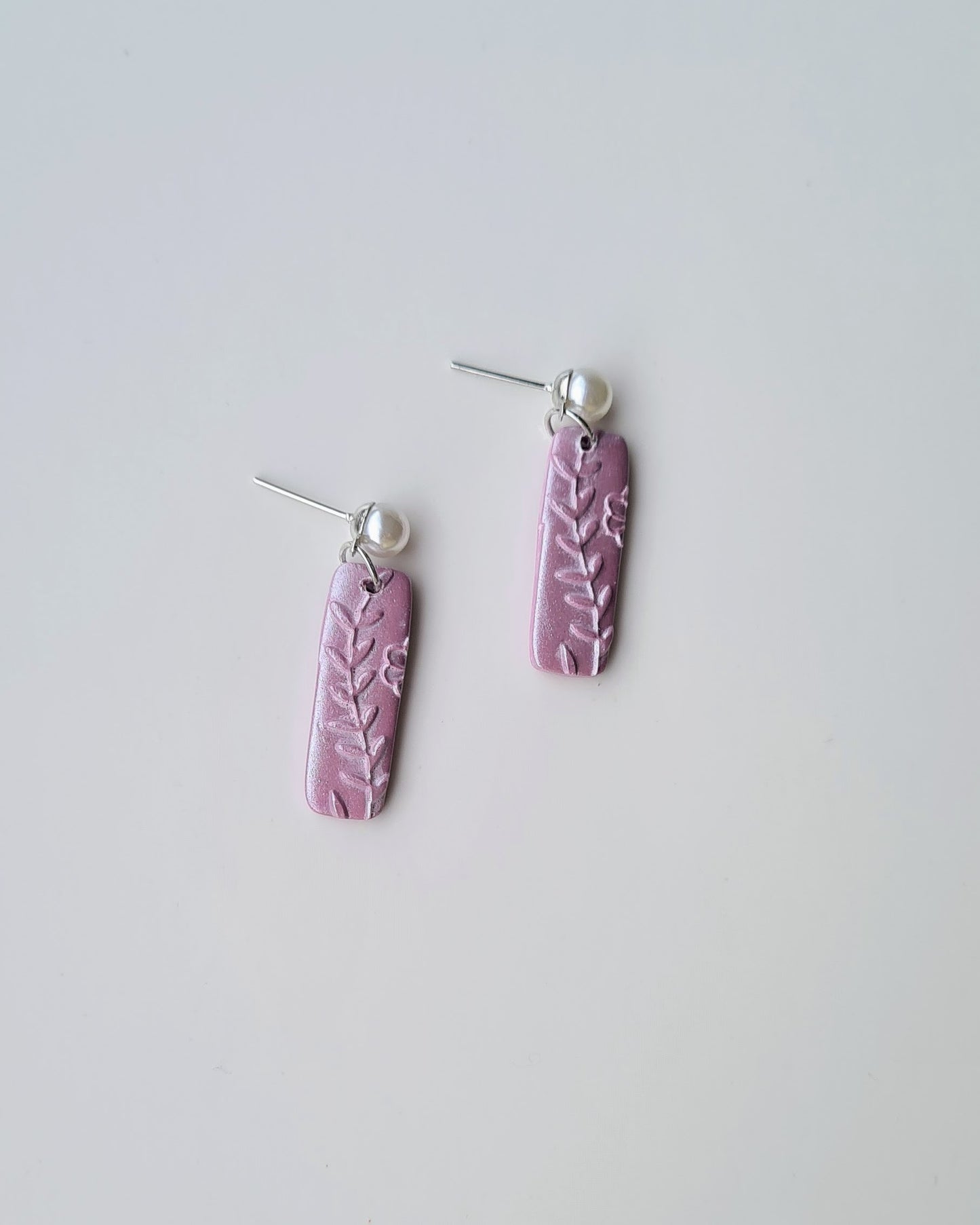 Pair of purple earrings with leaf patterns and pearl pendants on a light grey background