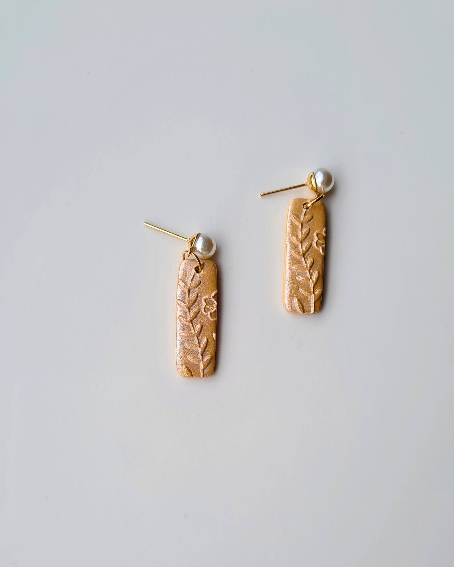Pair of shimmery yellow earrings with floral patterns and pearl pendant on a light grey background