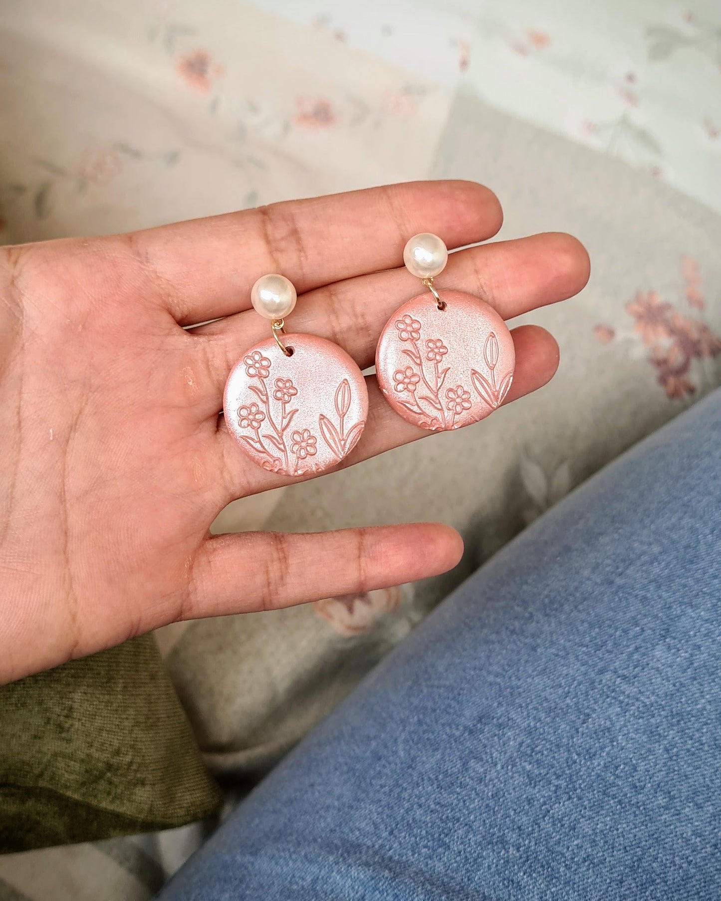 Hand holding a pair of earrings with floral designs on a patterned fabric background