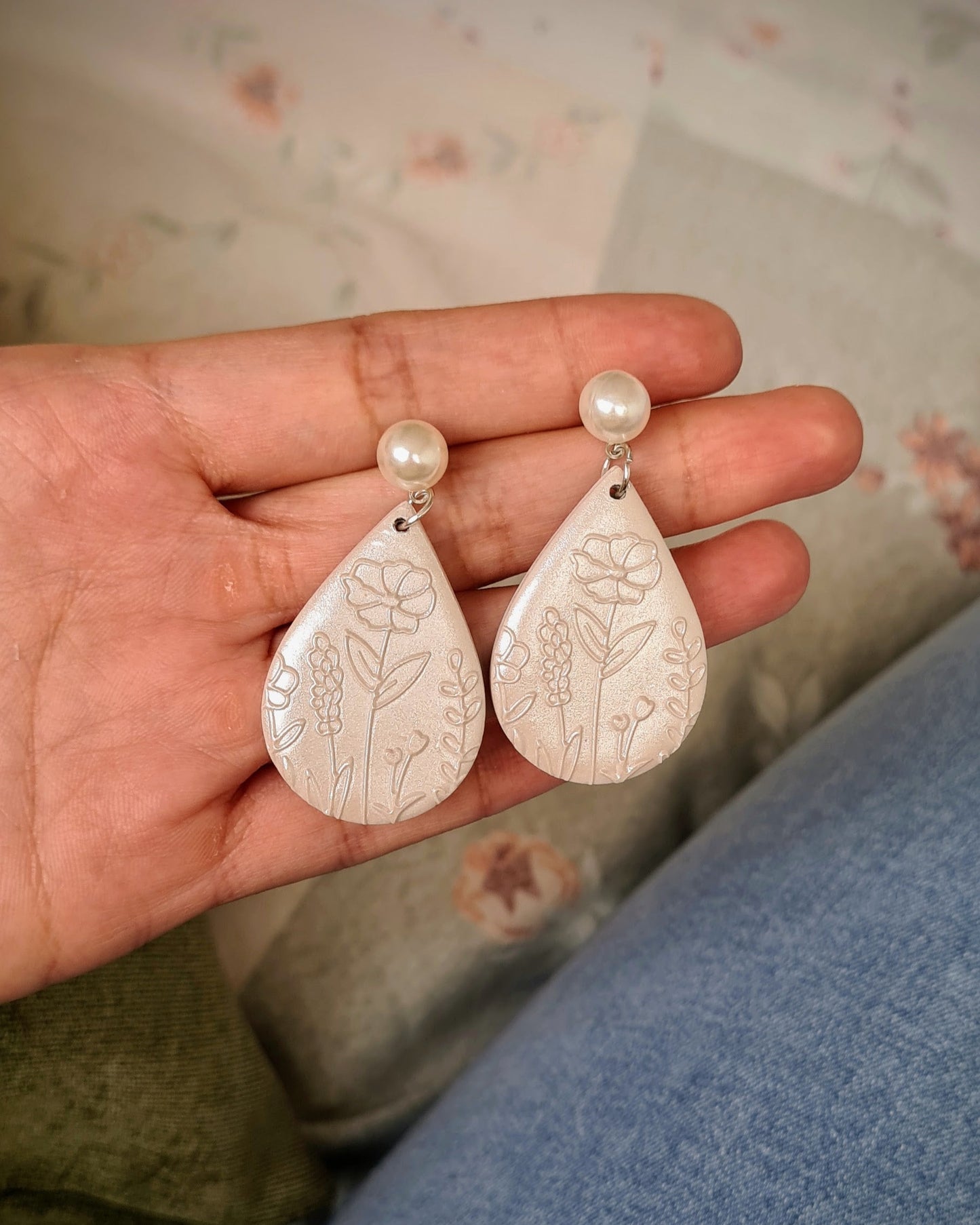 Teardrop-shaped pearlescent earrings with pearls held in a hand against a floral-patterned background.