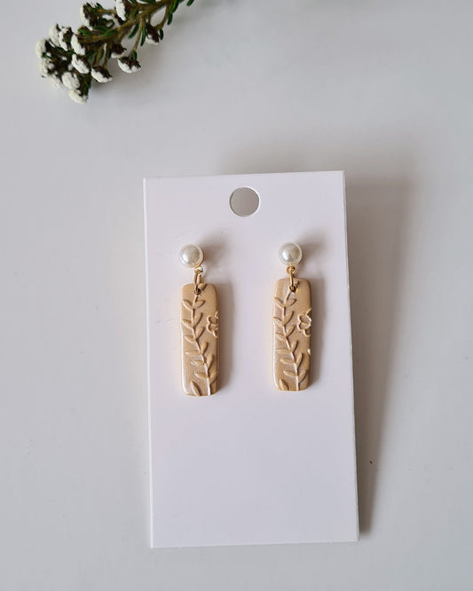 Pair of yellow shimmery earrings with pearl accent on a white card with greenery around