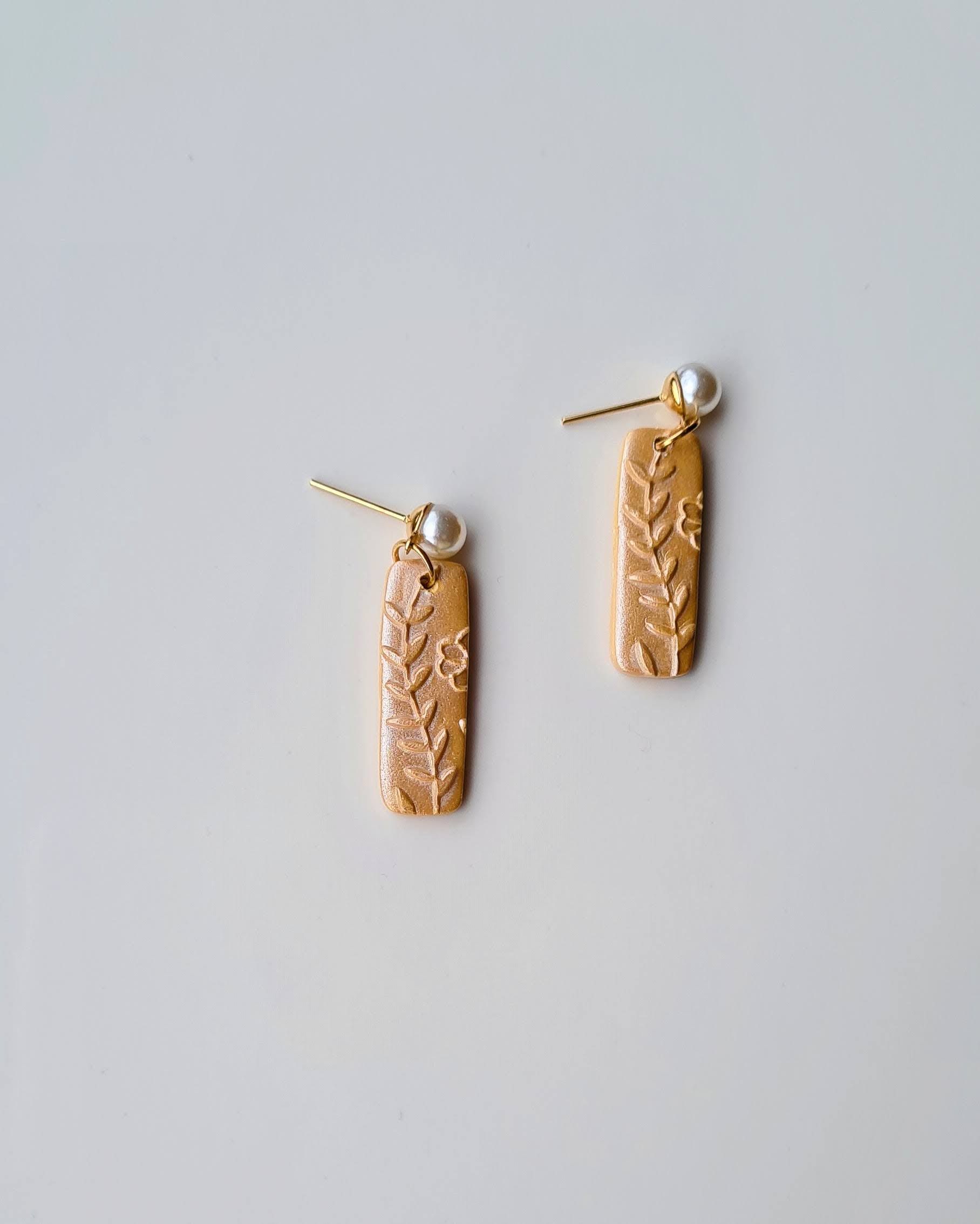 Pair of shimmery yellow earrings with floral patterns and pearl pendant on a light grey background