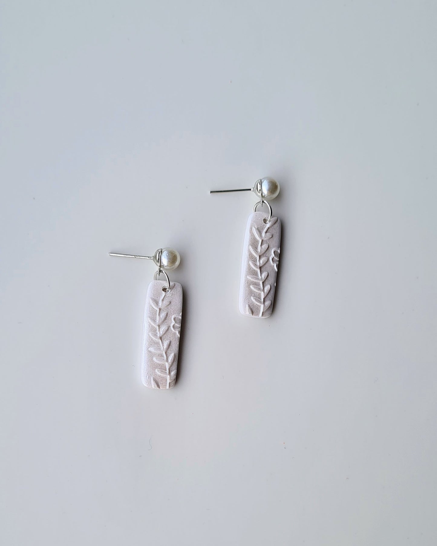 Pair of shimmery white earrings with leaf patterns and pearl pendant on a light grey background