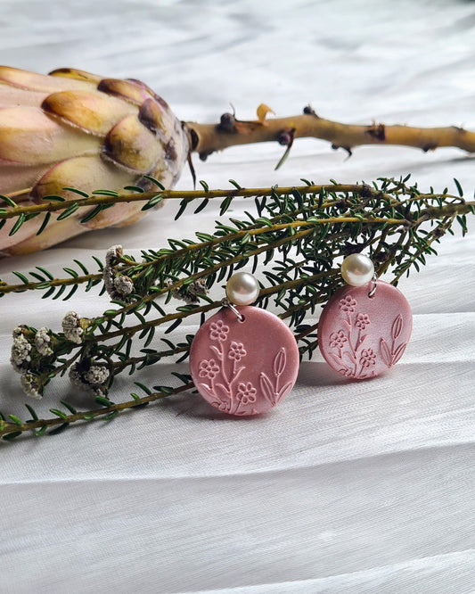 Pink shimmery earrings on a satin material surface with greenery