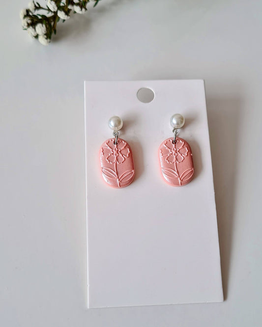 Pink oval-shaped earrings with floral texture and pearl accent on a white card with greenery around