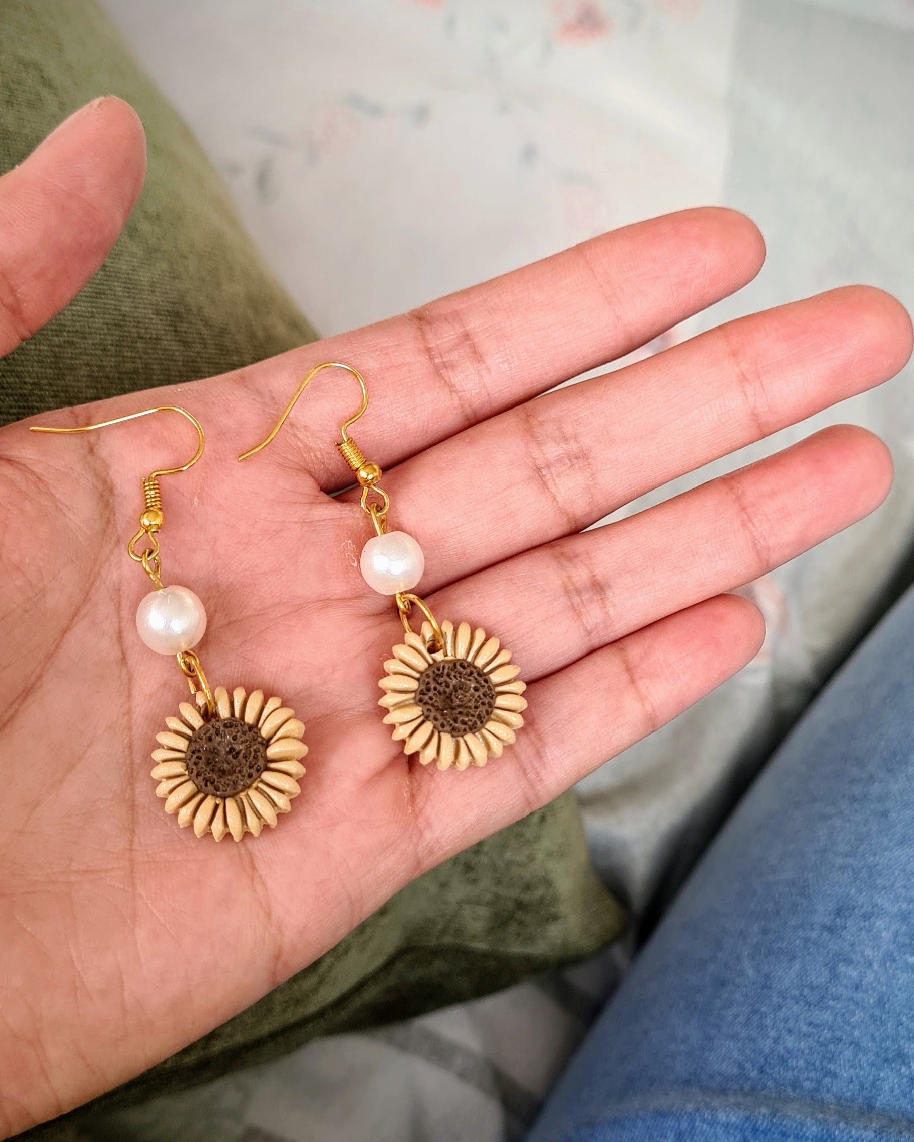 Hand holding two gold sunflower-shaped earrings with pearl centers on a light background.